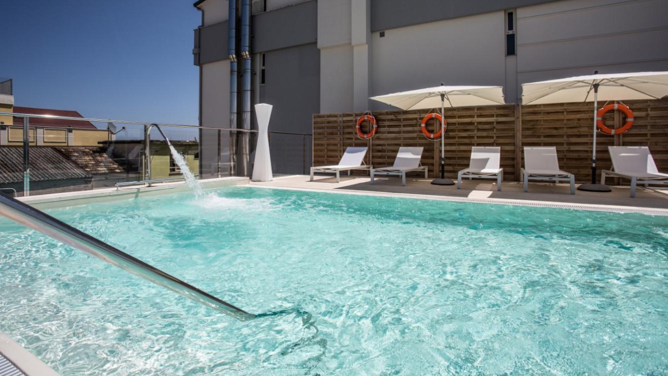 Piscina all'aperto con lettini, ombrelloni e salvagenti su una terrazza soleggiata.