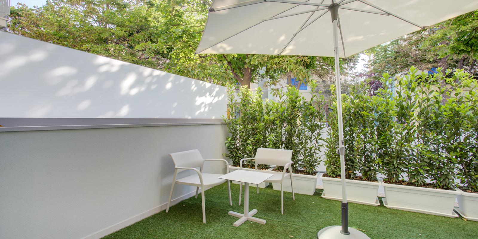 Terrazza con sedie bianche, tavolino e ombrellone su prato sintetico.