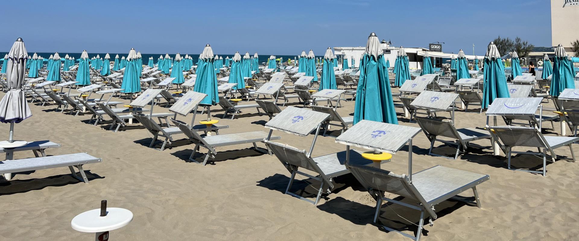 Spiaggia con lettini e ombrelloni chiusi, cielo sereno e mare sullo sfondo.