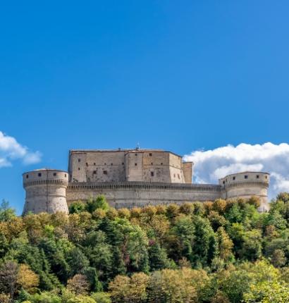 Imponente fortezza medievale circondata da alberi, sotto un cielo blu e nuvole bianche.