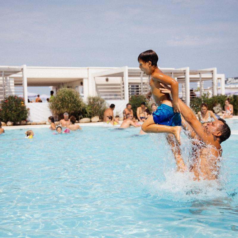 Papà solleva il figlio in piscina, circondati da bagnanti felici.