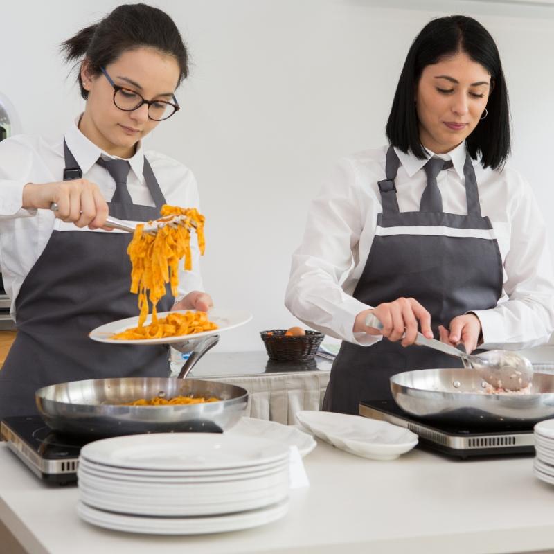 Due cuoche servono pasta in una cucina professionale con grembiuli grigi.