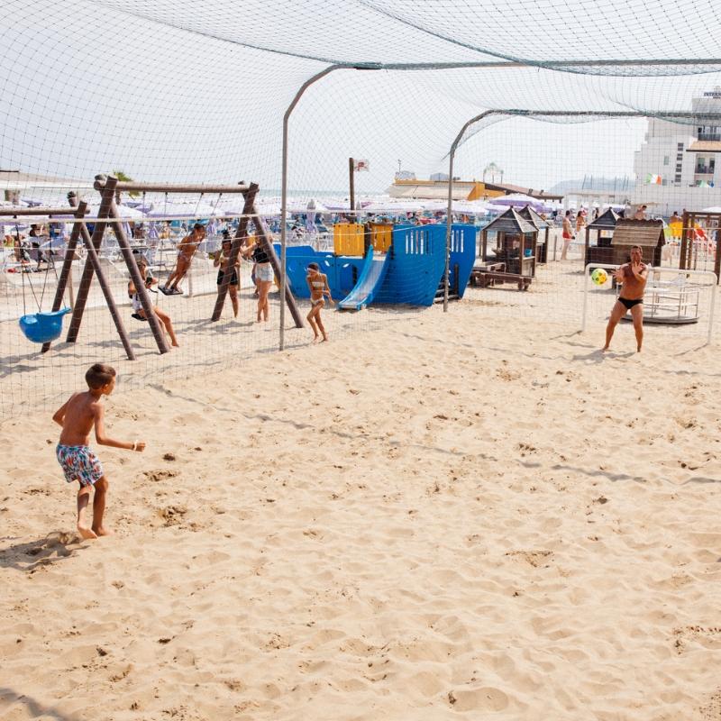 Spiaggia con area giochi per bambini, rete di protezione e persone che giocano a pallone.